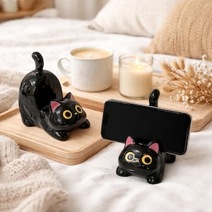 May include: Two black cat-shaped phone holders with yellow eyes and pink ears. One cat holds a phone, the other is empty. They sit on a wooden tray with a mug and a candle.