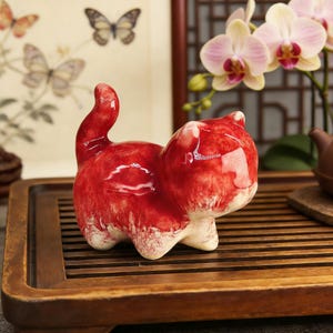 Puede incluir: Figura de gato de cerámica roja brillante con detalles blancos. El gato está representado en una pose juguetona, con la cola curvada hacia arriba. Se encuentra sobre una bandeja de madera, lo que sugiere un objeto decorativo o de colección.