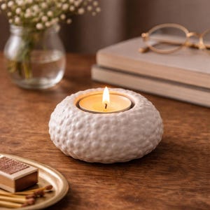 Pode incluir: Uma vela acesa em um porta-velas de cerâmica branca texturizada. O porta-velas tem formato redondo com superfície com covinhas. A chama da vela é brilhante contra os tons suaves e neutros do porta-velas e da decoração.