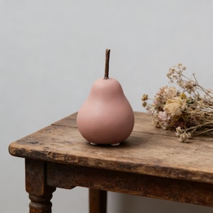 May include: A pink pear-shaped ornament with a brown stem. The pear sits on a rustic wooden table. Dried flowers are to the right of the pear. The pear has three small white feet.