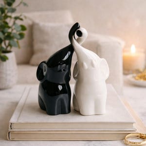 May include: A pair of ceramic elephant figurines, one black and one white, with intertwined trunks. The elephants are positioned on top of a stack of books. The background includes a lit candle and a plant in a white pot.