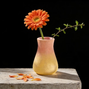 Puede incluir: Un pequeño jarrón degradado en tonos melocotón y amarillo contiene una gerbera naranja y una ramita de vegetación. El jarrón está sobre una superficie de hormigón, con pétalos esparcidos. El fondo es negro. Arreglo floral decorativo.