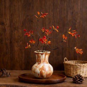 May include: A ceramic vase with a brown and white mottled design holds branches with vibrant orange and red leaves. The vase sits on a wooden tray, with a woven basket and pinecone in the background, creating a rustic, autumnal aesthetic.