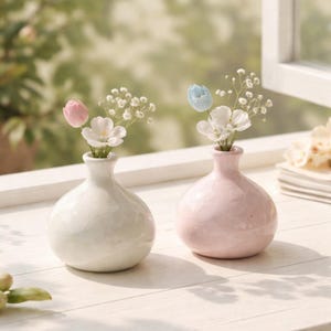 May include: Two small, round ceramic vases, one white and one pink, each holding delicate flowers. The white vase has a pink tulip and white flowers, while the pink vase has a blue tulip and white flowers. The vases sit on a white surface.