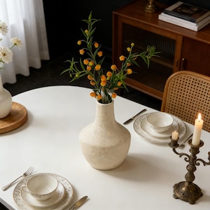 May include: A cream-coloured speckled vase holds orange and green floral stems, set on a white table. The table is set with a teacup, saucer, and plate with a gold floral design. A lit candle sits in a bronze candelabra.