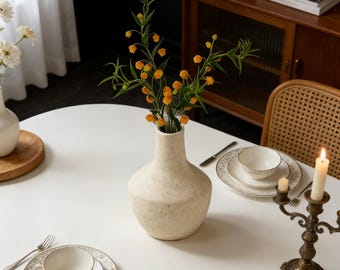 Pode incluir: Um vaso creme salpicado contém caules florais laranja e verdes, colocado sobre uma mesa branca. A mesa está posta com uma chávena, pires e prato com um desenho floral dourado. Uma vela acesa está num castiçal de bronze.