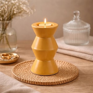 May include: A yellow ceramic candle holder with a lit tea light candle. The holder has a unique hourglass shape and sits on a round woven placemat. A glass jar and vase with flowers are in the background.