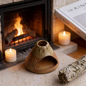 May include: A volcano-shaped ceramic essential oil diffuser, glazed in brown and green. It's placed on a stone surface next to a lit candle, a bundle of sage, and a fireplace with a burning fire.