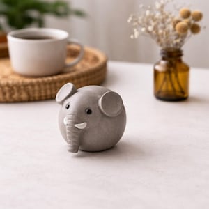 May include: A grey ceramic elephant ornament with a rounded body, small ears, and a trunk. The elephant has black eyes and white tusks. The ornament is on a white surface, with a cup of coffee and a decorative vase in the background.