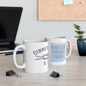 Puede incluir: Dos tazas de cerámica blanca. Una taza tiene el texto "Gibbs' R" con un diseño de nudo náutico y "N. 1" en azul marino. La otra taza tiene el texto "Never Screw Over Your Partner" en una nota adhesiva azul claro.
