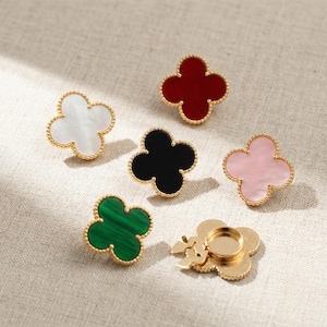 May include: A collection of clover-shaped jewelry pieces in various colors, including white, red, black, pink, and green. Each piece has a gold-colored beaded border. One piece has a clasp on the back.
