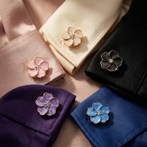Puede incluir: Primer plano de gemelos con forma de flor en varios colores: rosa, crema, negro, morado y azul. Cada gemelo tiene un borde dorado y una pequeña gema brillante en el centro. Los gemelos se muestran en los puños doblados de camisas.