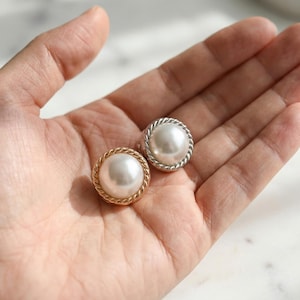 以下が含まれることがあります： ねじれたロープのデザインのボーダーが付いた、2つの丸い真珠のイヤリング。片方のイヤリングはゴールドトーンのボーダー、もう片方はシルバートーンのボーダーです。イヤリングは明るい色の表面に表示されています。