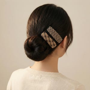 Könnte beinhalten: Zwei rechteckige Haarspangen mit Hahnentrittmuster in Braun- und Beigetönen sind im Haar platziert. Das Haar ist zu einem tiefen Dutt gebunden. Die Person trägt einen cremefarbenen Pullover.