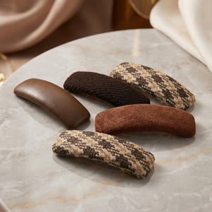 Könnte beinhalten: Eine Auswahl an Haarspangen in verschiedenen Brauntönen. Die Spangen weisen unterschiedliche Texturen auf, darunter glattes Leder, gewebter Stoff und ein tweedartiges Muster. Die Haarspangen sind auf einer Marmoroberfläche angeordnet.