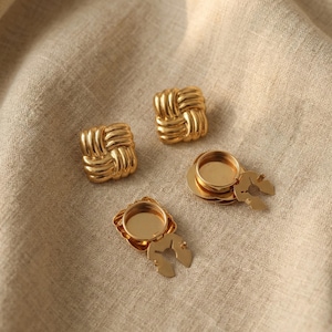 Puede incluir: Cuatro componentes de joyería dorados sobre una tela beige. Dos son tachuelas cuadradas de estilo tejido. Los otros dos son redondos con un cierre decorativo en forma de hoja. Los componentes son probablemente para pendientes u otras joyas.