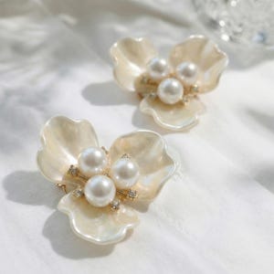 May include: Two flower-shaped brooches with creamy white petals and three large, round pearls at the centre. Small, clear crystals are set around the pearls. The brooches are on a white background.
