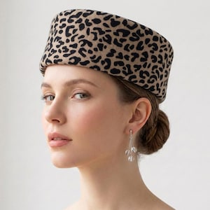 Op de afbeelding: Een hoed met luipaardprint in beige en zwart. De hoed is een pillbox-stijl, met een platte bovenkant en een afgeronde rand. De hoed wordt gedragen door een persoon met een lichte huid, bruin haar en zilveren oorbellen. De oorbellen hebben drie heldere, ronde kralen.