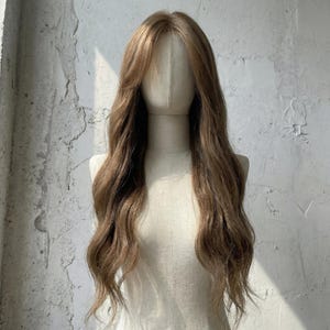 Puede incluir: Una peluca larga y ondulada en tonos marrones y rubios se exhibe en un maniquí. La peluca presenta una línea de cabello de aspecto natural y mechones largos y fluidos. El cabello está peinado con ondas suaves y una raya lateral.