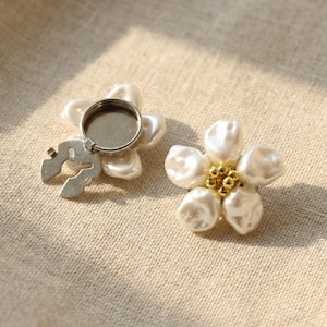 Puede incluir: Dos broches con forma de flor. Cada broche presenta cinco perlas de imitación blancas de forma irregular. Un broche tiene una base de metal plateado con un pequeño lazo y un engaste circular en blanco. El otro broche tiene un grupo de pequeñas cuentas doradas en el centro.