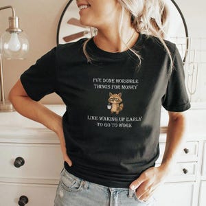Puede incluir: Camiseta negra con el texto "I'VE DONE HORRIBLE THINGS FOR MONEY" y un mapache de dibujos animados sosteniendo una taza de café. Debajo, el texto "LIKE WAKING UP EARLY TO GO TO WORK".