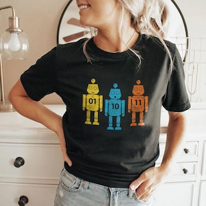 Peut inclure: T-shirt noir avec trois illustrations de robots rétro en jaune, bleu et orange. Chaque robot porte un numéro : 01, 10 et 11. Le design a un aspect vieilli et vintage.