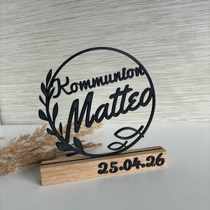 May include: A black laser-cut Communion table decoration. The design features the word "Kommunion Matteo" within a circular frame, adorned with leaves. The date "25.04.26" is displayed on a wooden base.
