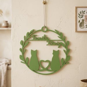 Könnte beinhalten: Ein kreisförmiger grüner Wandbehang mit einem blättrigen Kranzrand, zwei voneinander abgewandten Katzensilhouetten und einer herzförmigen Aussparung in der Mitte. Der Schriftzug "Familie König" ist im oberen Bereich ausgeschnitten.