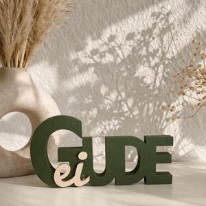 Op de afbeelding: Een donkergroen, driedimensionaal decoratief bord met het woord Gude, waarbij de letters ei in een lichtere natuurlijke houttint aan de voorzijde van de letter G zijn bevestigd. De letters zijn blokvormig en staan rechtop.