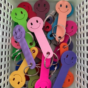 Op de afbeelding: Een verzameling veelkleurige sleutelhangers, elk met een cirkelvormig smiley-embleem aan de bovenkant en een lange, platte rechthoekige steel. De sleutelhangers zijn gemaakt van effen gekleurd plastic en bevatten bevestigde zilverkleurige metalen ringen.
