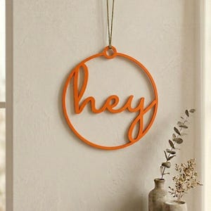May include: A bright orange circular wall hanging featuring the word hey in a cursive script font. The piece is suspended by a thin cord from a small wooden hook. The design is constructed from a single continuous frame with integrated lettering.