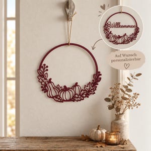 Pode incluir: Um adorno de parede circular vermelho escuro com um design recortado de abóboras e folhas de outono. Um círculo menor mostra o mesmo design com o texto Willkommen. Um balão de texto indica Auf Wunsch personalisierbar.