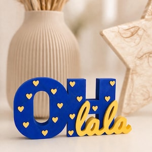 Könnte beinhalten: Blaues Holzschild mit den Worten "OH lala" in Gelb. Die Buchstaben "OH" und "H" sind blau mit gelben Herzakzenten. Das Wort "lala" ist in gelber Kursivschrift geschrieben.