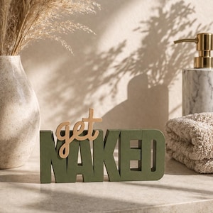 Könnte beinhalten: Dekoratives Holzschild mit dem Schriftzug "get NAKED". Das Wort "NAKED" ist in Dunkelgrün gehalten, während "get" in einem helleren Holzton gehalten ist. Das Schild steht auf einer hellen Oberfläche.