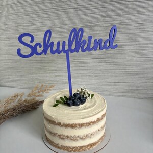 Op de afbeelding: Een ronde, gelaagde cake met witte glazuur en een blauwe "Schulkind" taarttopper. De cake is versierd met blauwe bessen, kleine witte bloemen en groene bladeren. De cake staat op een heldere taartplank.