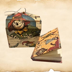 May include: A decorative, vintage-style box and small book. The box features a smiling scarecrow illustration with a red hat and green clothing. The book has a yellow cover with illustrations and open pages.