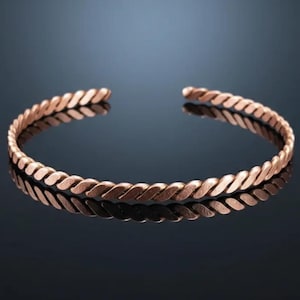 Puede incluir: Una pulsera de puño abierta de color cobre con un diseño de cuerda trenzada. La pulsera está hecha de un metal brillante y refleja la luz. La joya se exhibe sobre una superficie oscura y reflectante.