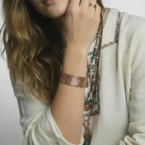 May include: A copper-colored cuff bracelet with an intricate floral design. The bracelet is worn on a person's arm, and the design is visible. The person is wearing a white shirt with colorful embroidery and multiple necklaces.