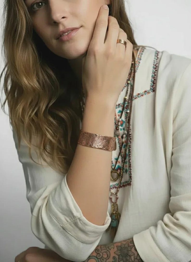 May include: A copper-colored cuff bracelet with an intricate floral design. The bracelet is worn on a person's arm, and the design is visible. The person is wearing a white shirt with colorful embroidery and multiple necklaces.
