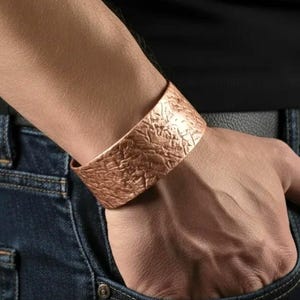 May include: A copper-colored cuff bracelet with a textured surface. The wide band bracelet is worn on a wrist, with the hand in a blue jeans pocket. The bracelet has a hammered or embossed appearance.