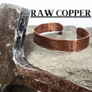 Puede incluir: Una pulsera de cobre con superficie texturizada está junto a un martillo. La pulsera es abierta y tiene un aspecto rústico y hecho a mano. El texto "RAW COPPER" está encima de la pulsera. El fondo es gris neutro.