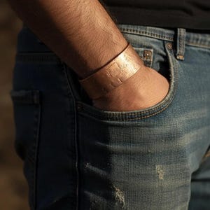 May include: A copper cuff bracelet with a textured surface is worn on a person's wrist. The bracelet is wide and appears to be handmade. The person is wearing blue jeans and a black shirt.