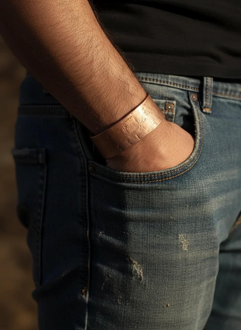 May include: A copper cuff bracelet with a textured surface is worn on a person's wrist. The bracelet is wide and appears to be handmade. The person is wearing blue jeans and a black shirt.