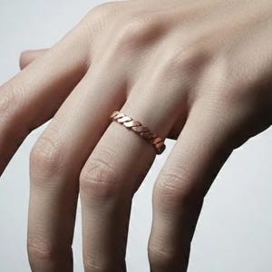 May include: A rose gold ring with a woven design, worn on a finger. The ring's intricate pattern is visible, with a warm metallic hue. The ring's design is a continuous braid, adding a touch of elegance. The ring's design is a continuous braid.