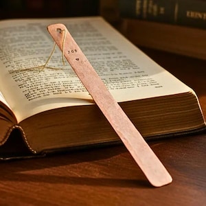 May include: A copper bookmark with the text "JOS" and a small heart shape, resting in an open book. The bookmark is rectangular with a small hole at the top and a thin string attached. The book has aged, yellowed pages.