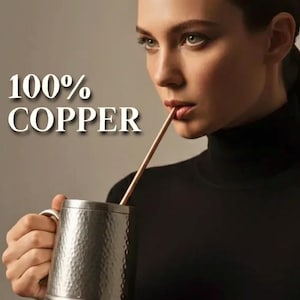 May include: A hammered copper mug with a copper straw. The mug has a handle and a flared base. The text "100% COPPER" is displayed in white. The person in the image is wearing a black turtleneck.