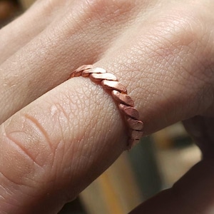 May include: A copper-coloured ring with a braided design. The ring is worn on a finger, showcasing its intricate woven pattern. The ring's warm hue and textured surface create a unique and stylish accessory.