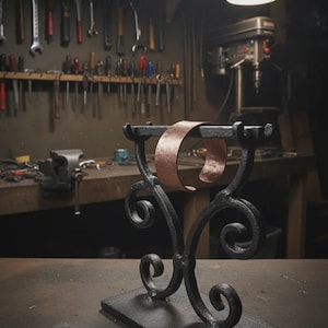May include: A handcrafted, black metal bracelet display stand with a copper bracelet. The stand features decorative scrollwork and a rectangular base. The workshop background includes tools and equipment.