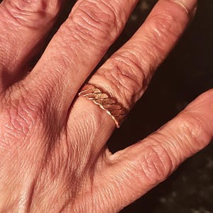 May include: A gold-coloured ring with a woven or braided design. The ring is worn on a finger, showcasing its intricate pattern and metallic sheen. The ring's design is a continuous loop, creating a textured surface.