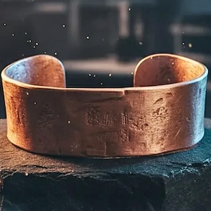 May include: A copper cuff bracelet with a textured surface. The open-ended bracelet has a rectangular shape and is displayed on a dark surface. The bracelet is a warm brown color.